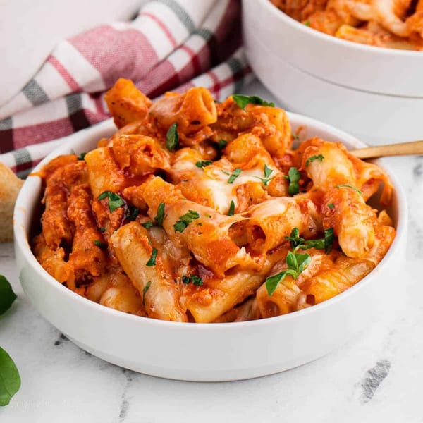 A white bowl filled with rigatoni pasta in a creamy red sauce, topped with melted cheese and fresh parsley.