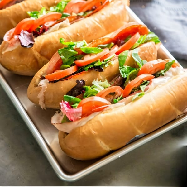 Four Casa Bianca subs with fresh tomatoes, lettuce, and sauce on a metal tray.