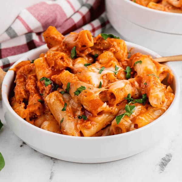 Close-up of a white bowl filled with rigatoni pasta in a creamy red sauce, topped with melted cheese and fresh parsley.