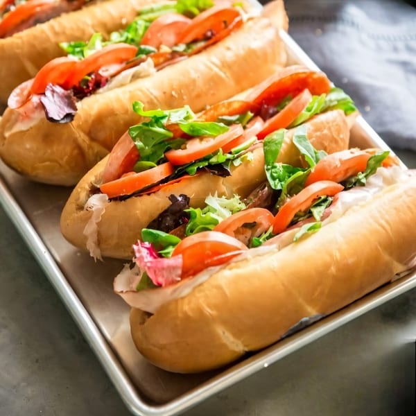 Four fresh subs with tomatoes, lettuce, and meat on a metal tray.